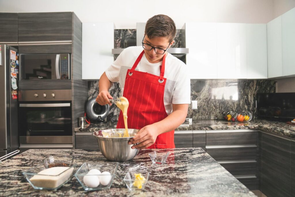 A teenaged boy wearing a red apron and stirring something in an aluminum bowl