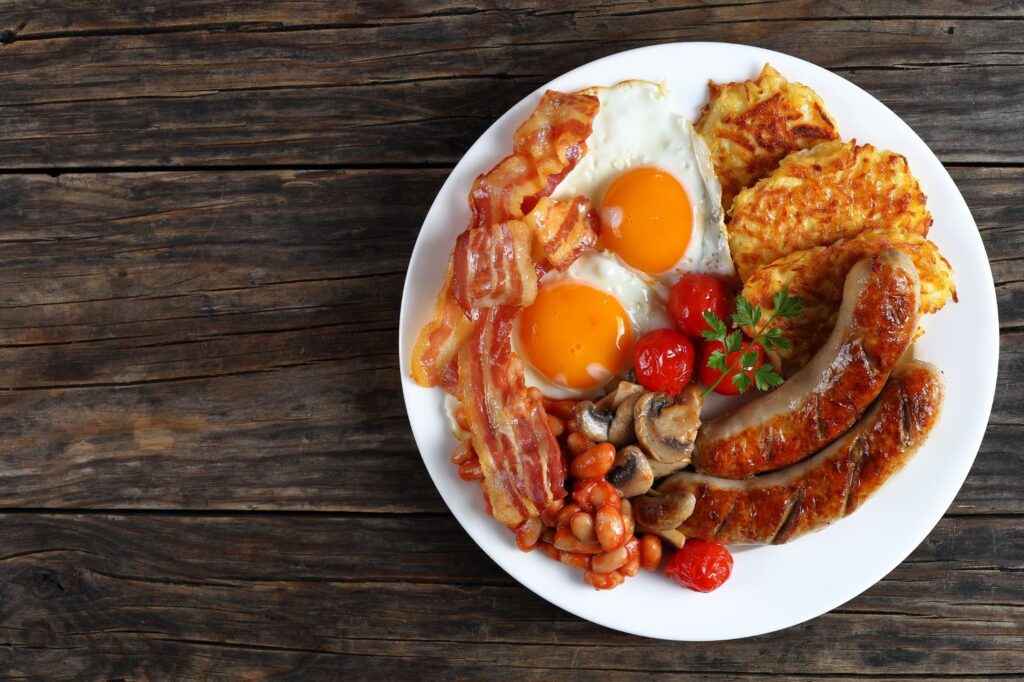 A plate with a traditional English breakfast with sausages, bacon, eggs, beans, tomatoes and hash browns. 