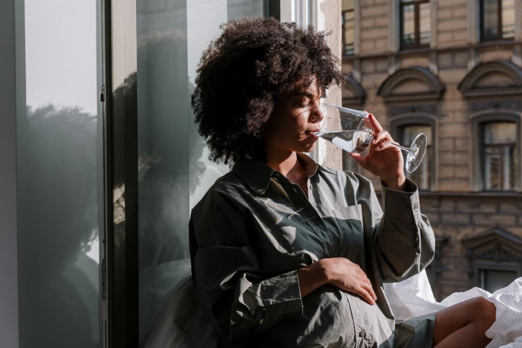 A pregnant woman drinking a glass of water while sitting beside a large window