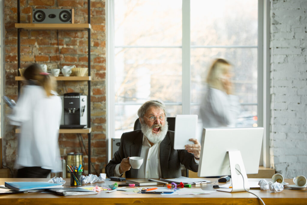 An angry boss yelling while seated at his desk as employees move quickly around him