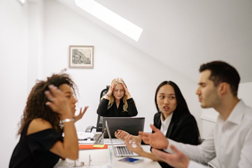A group of employees arguing with each other while a woman in the back of the room holds her head in frustration