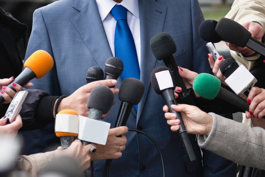 A man in a suit trying to play himself up while talking to reporters, who are holding up microphones and recording devices