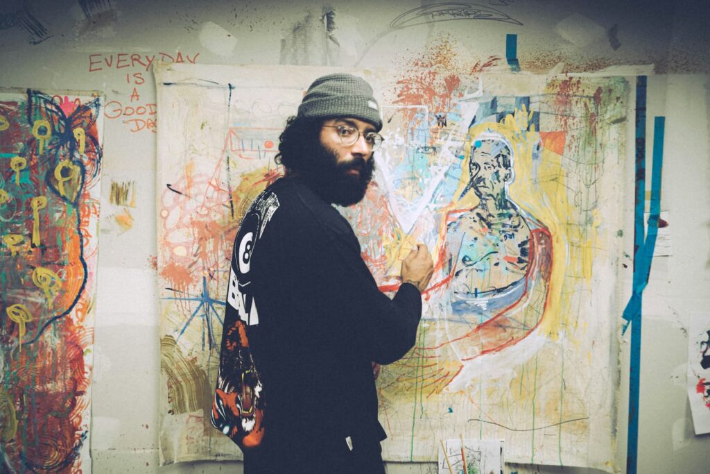A young man in a hat making an abstract painting a canvas hanging on a wall