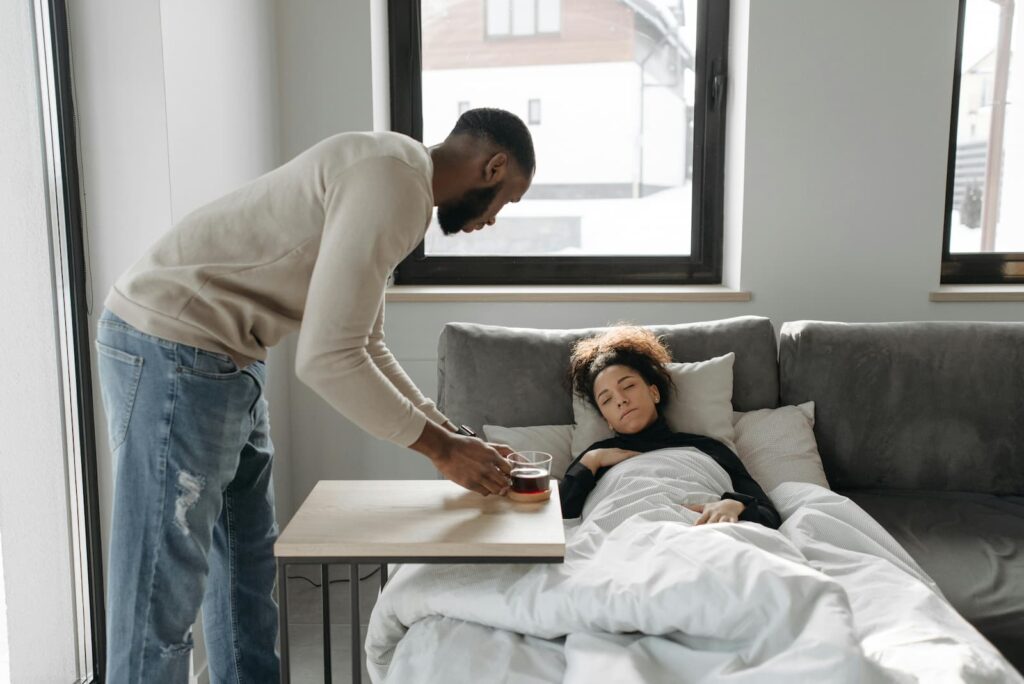 A man bringing tea to his sick girlfriend, who is lying under a blanket on a sofa