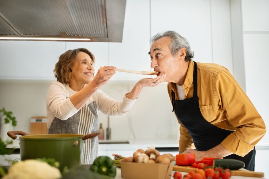 A woman in an apron holding out a spoon while a man sips from it
