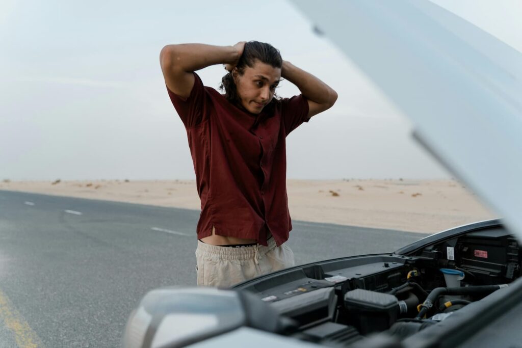 A frustrated man looking under the hood of his car that is stopped on the side of a road