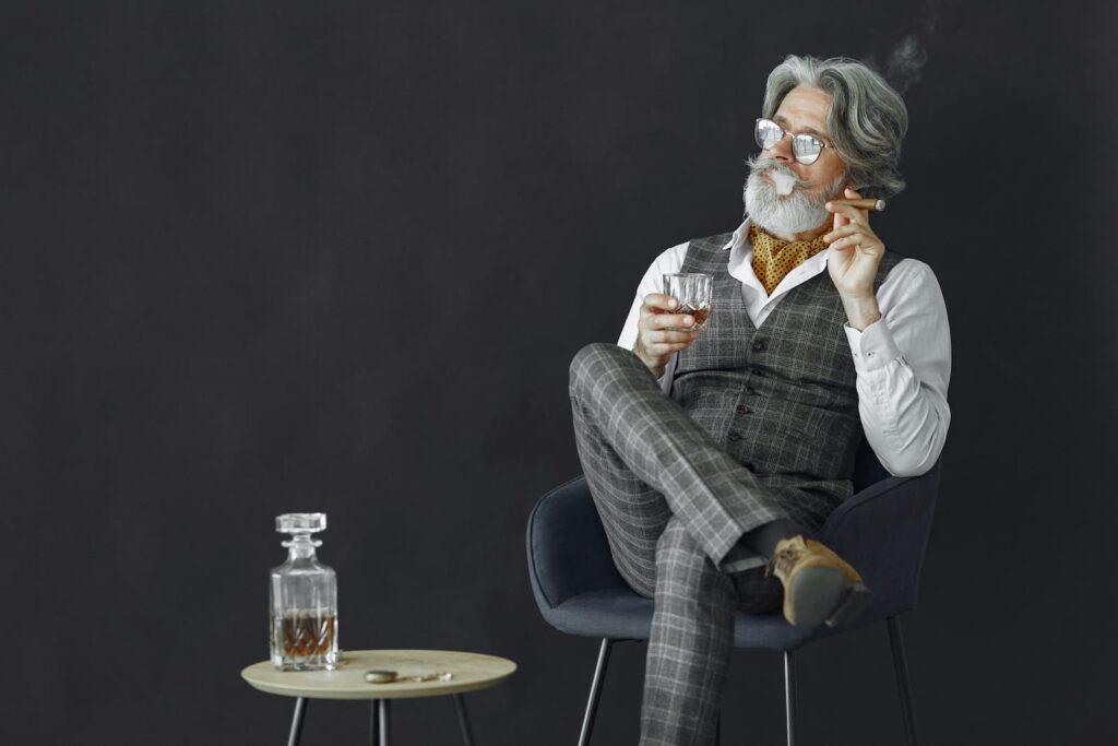 A well-dressed older man holding a cigar in one hand and a glass of alcohol in the other