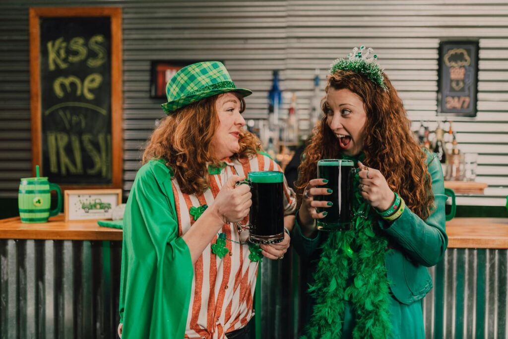 Two women wearing green holding large mugs of dark beer while standing in a bar